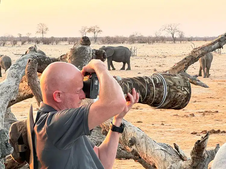 A man photographing wildlife with a DSLR camera and large camouflage telephoto lens, framed by dry tree branches in an African savanna landscape. Several elephants roam in the background under a warm, golden sky on an African photo safari.