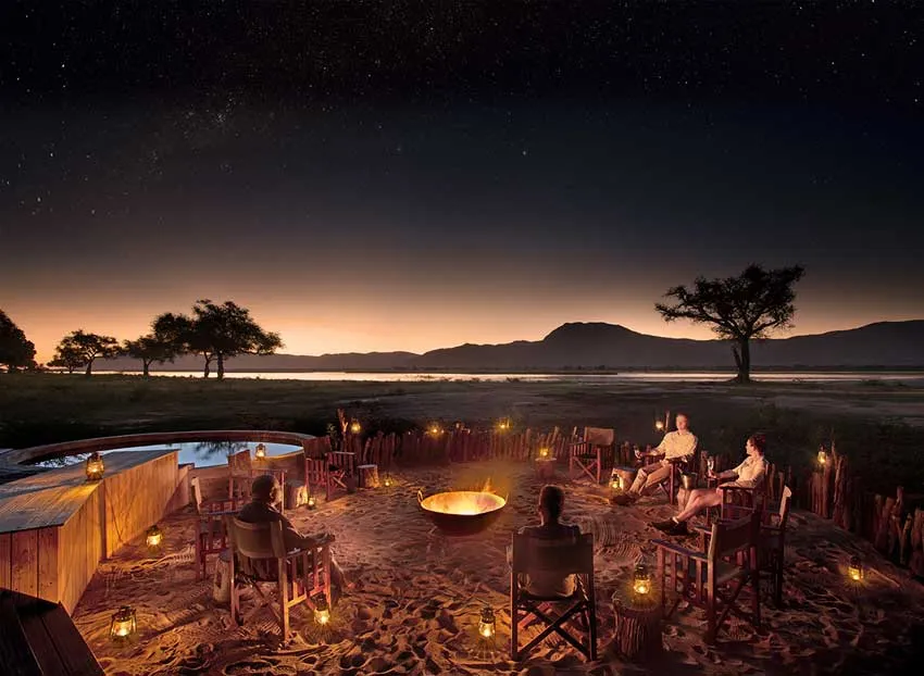 An African safari experience at Nyamatusi Mahogany Camp by African Bush Camps in Mana Pools, where guests sit around a lantern-lit firepit with views of the wilderness and starry skies.
