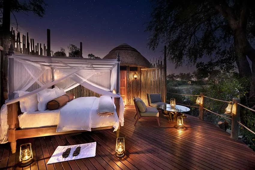 Luxury African safari lodge with a canopy bed on a private deck under the stars, surrounded by lantern light and natural bush setting.