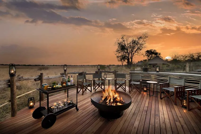 A cozy firepit and outdoor lounge area at Khwai Lediba Lodge in Botswana, part of African Bush Camps. Set against a golden sunset over the open floodplains, this luxury African safari setting features director’s chairs, lanterns, and a bar cart ready for sundowners.