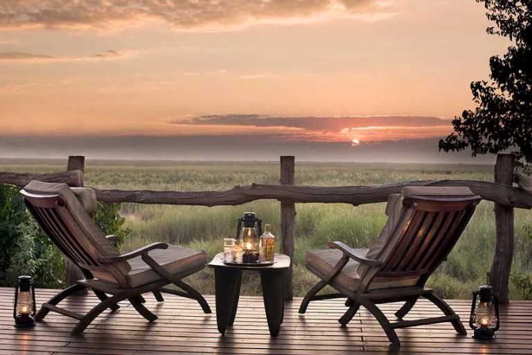 Sunset at Linyanti Bush Camp by African Bush Camps, Botswana. Two safari chairs on a wooden deck overlook misty floodplains, with a table set for sundowners—lanterns glowing, whisky and glasses ready after a day on safari.