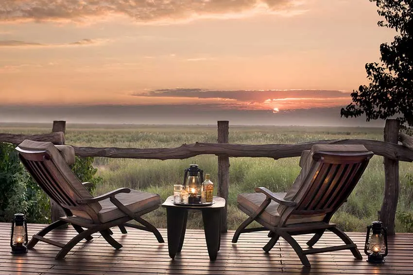 Sunset at Linyanti Bush Camp by African Bush Camps, Botswana. Two safari chairs on a wooden deck overlook misty floodplains, with a table set for sundowners—lanterns glowing, whisky and glasses ready after a day on safari.