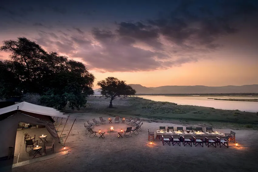 Scenic Mana Pools African safari camp overlooking the river at sunset, with a glowing campfire circle and lantern-lit dining table set for guests.