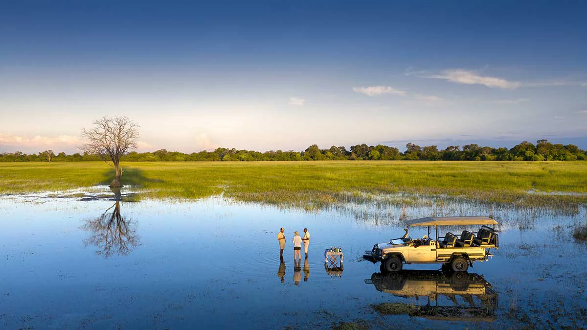 Three people enjoy sundowners beside a safari vehicle in the shallow floodplains of the Okavango Delta, Botswana. The peaceful scene features still water reflecting the sky and a lone tree, with lush green grasslands and clear skies creating a breathtaking African safari moment.