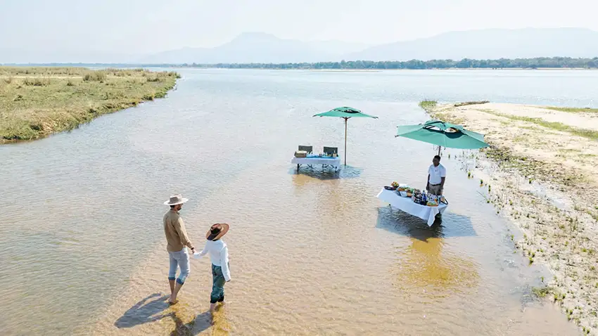 Couple enjoying a romantic riverside lunch setup in the Lower Zambezi, Zambia, with a private dining table and umbrellas set in shallow water during an African safari.