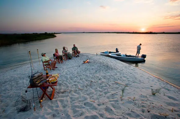 A small group relaxes with drinks and a campfire on a sandy riverbank in the Caprivi Strip, Namibia, with a fishing boat nearby and a stunning sunset over the water.