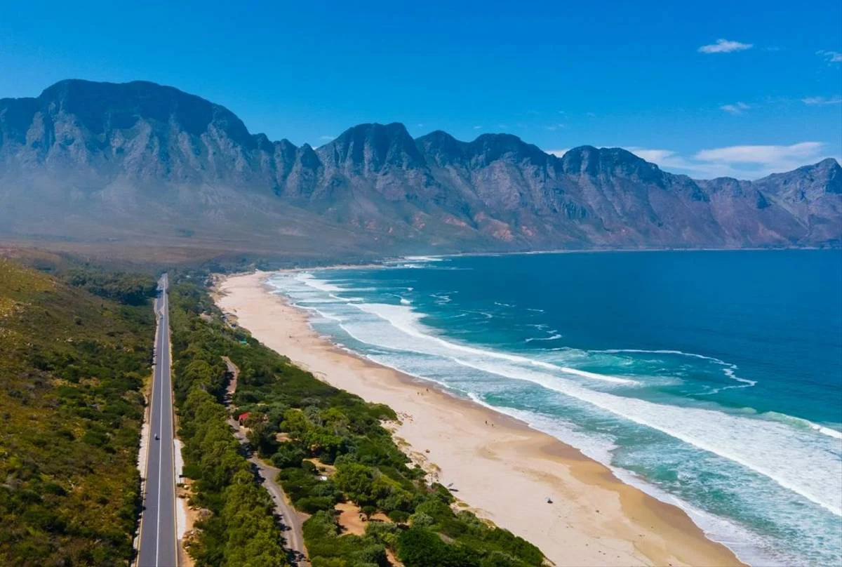 A scenic aerial view of South Africa’s Garden Route, showcasing a winding coastal road alongside a pristine beach and the dramatic backdrop of the Tsitsikamma Mountains. Crystal-clear waves roll onto the shore under a bright blue sky, capturing the beauty of this iconic road trip destination.