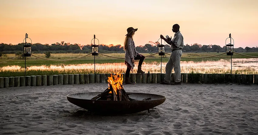 A guest and guide share stories by the firepit at a Moremi Game Reserve safari camp in Botswana. The sun sets over the wetland landscape, casting warm hues across the sky while lanterns and natural wood accents add to the intimate bush setting of this authentic African safari experience.