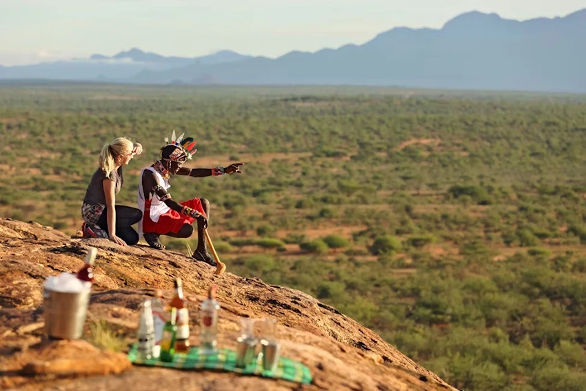 A Samburu warrior in traditional attire points across the vast landscape of northern Kenya, guiding a female guest seated beside him on a rocky outcrop. A picnic with drinks sits nearby, with the green valleys and distant mountains of the Samburu region stretching into the horizon—an authentic cultural experience on an African safari.