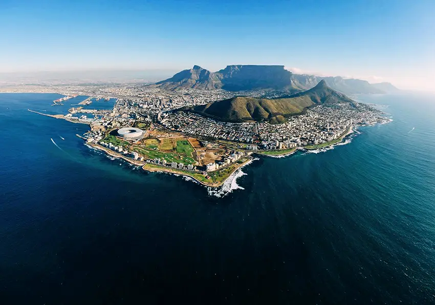 Aerial view of Cape Town, South Africa, with Table Mountain towering over the city, the Cape Town Stadium in the foreground, and the Atlantic Ocean surrounding the coastline. A stunning blend of natural beauty and urban landscape at the tip of the continent.