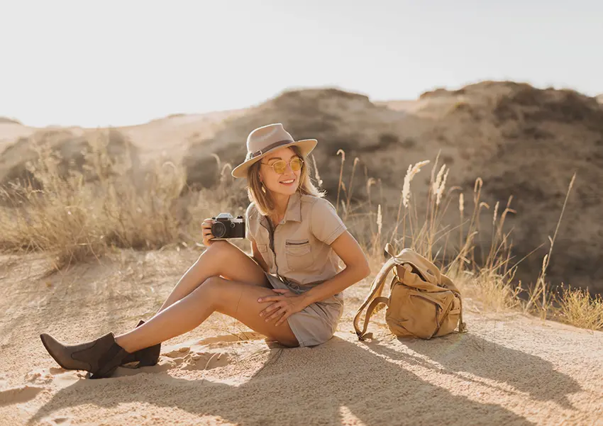 Smiling woman sitting on a sandy hill during an African photo safari, holding a camera with a backpack by her side, dressed in safari attire and sun hat.