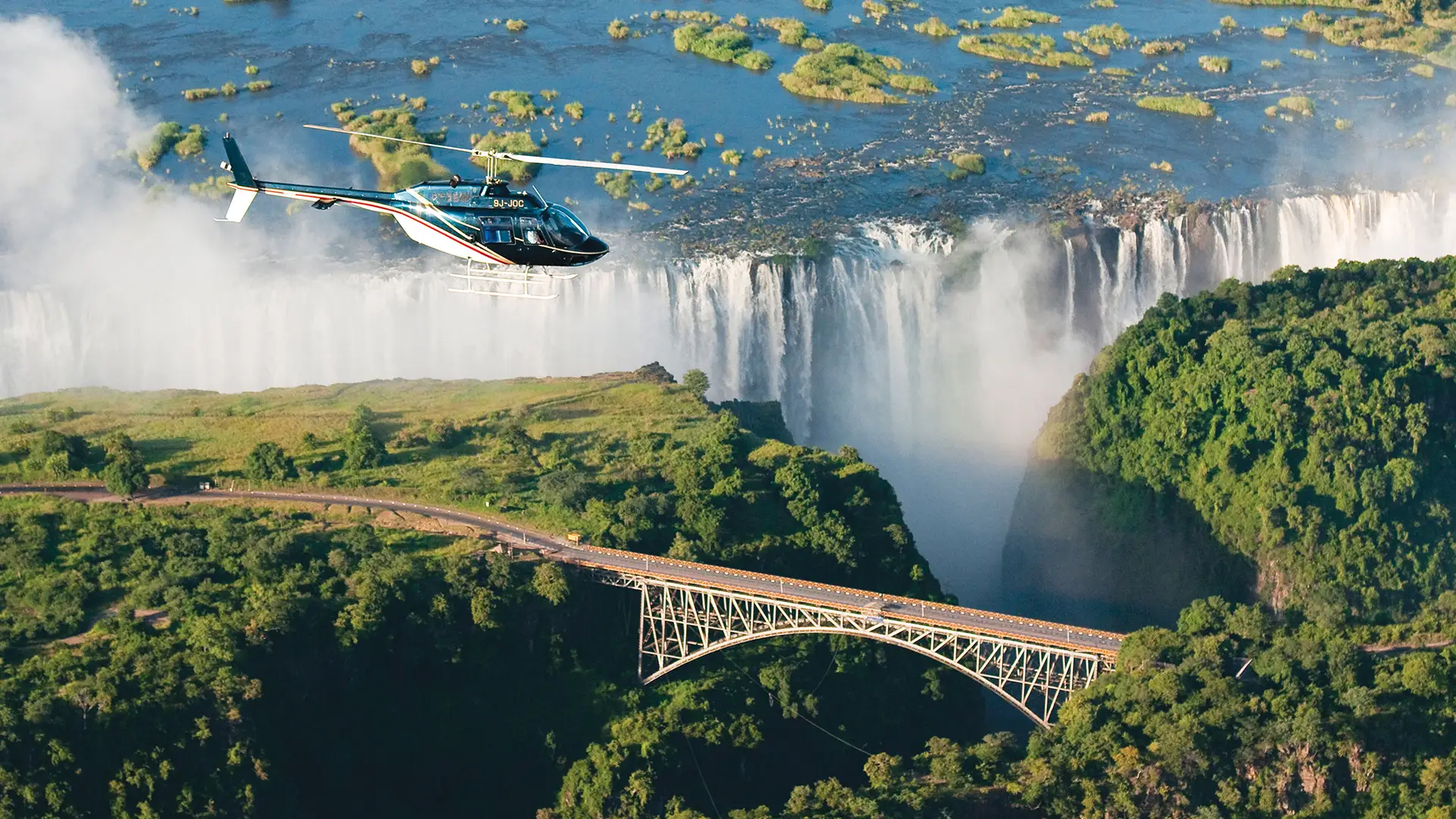A helicopter soars above Victoria Falls in Zimbabwe, with a bridge visible near the cascading waterfall below.