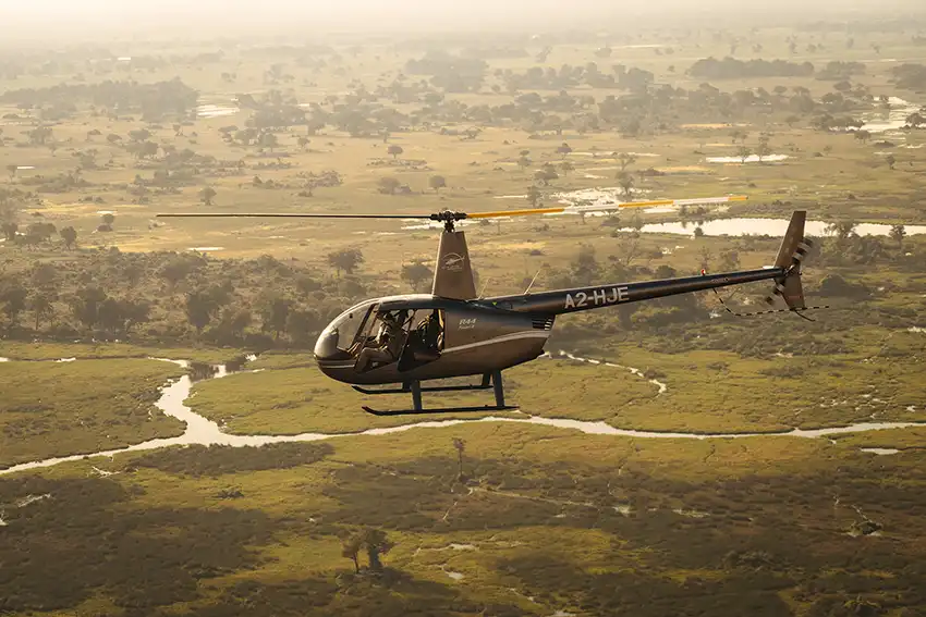A dark grey Helicopter Horizons helicopter flies over the Okavango Delta in Botswana, offering an aerial safari tour. The aircraft's blades spin against the golden light, showcasing the winding waterways and lush green landscape below.