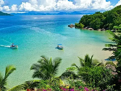 Tropical paradise with turquoise water, lush green hills, and two boats gently floating. White sand beach and palm trees complete the idyllic scene.