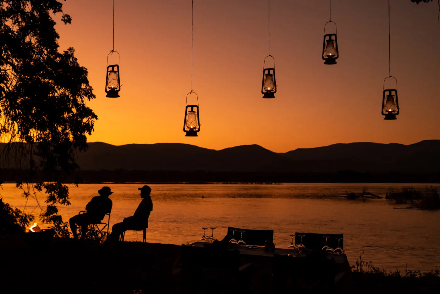 Silhouettes of two people sitting by a lake at sunset, with hanging lanterns and mountains in the background, evoke a peaceful atmosphere for Mana Pools safaris.