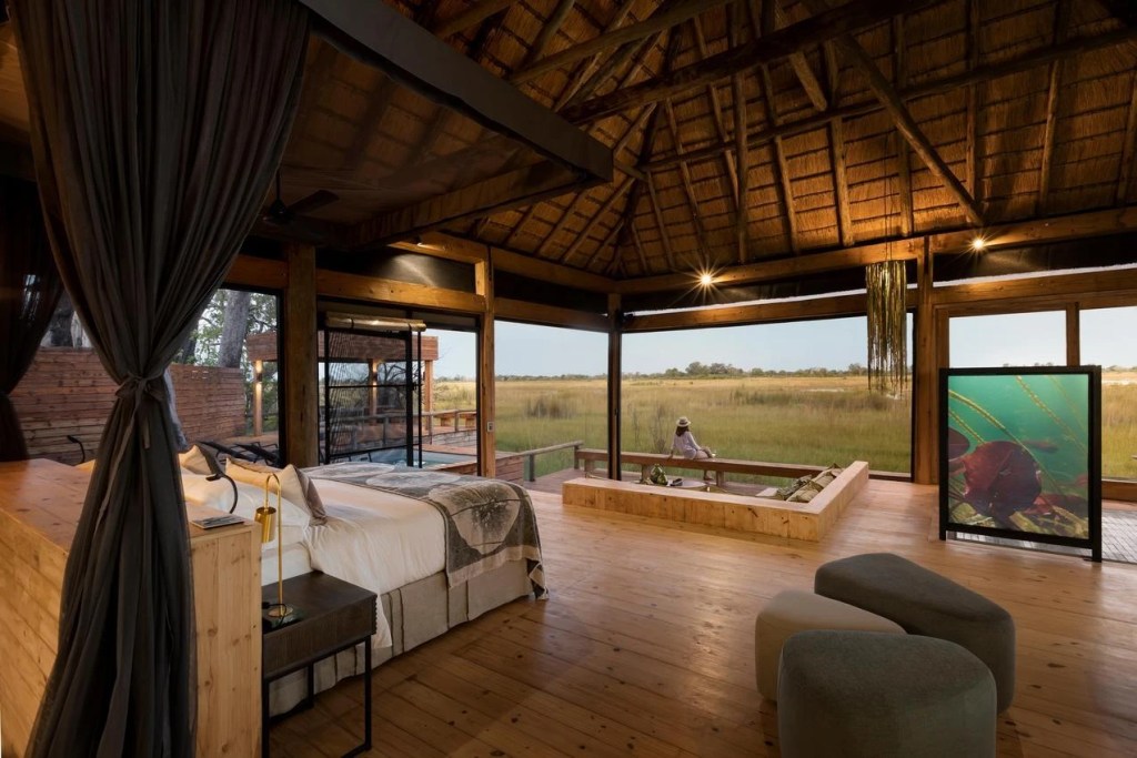 Luxury safari lodge bedroom with thatched roof, king-size bed, and large windows overlooking a vast African wetland landscape.